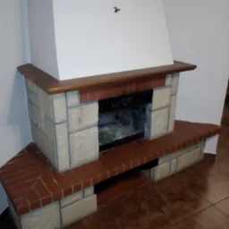 Otynkowany kominek z ceglaną i kamienną obudową, wykończony drewnianym blatem, na brązowej podłodze.