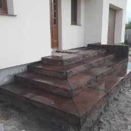 Granitowe schody wejściowe do domu, brązowo-czarna kostka, widoczne ślady zaprawy, boczny widok, częściowo ukończone, surowe otoczenie.