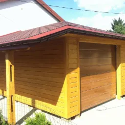 Drewniana altana z czerwoną dachówką, widoczna z zewnątrz, z elementami konstrukcyjnymi i roślinnością w tle. Jasne drewno, poziome deski.