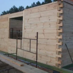 Surowy szkielet drewnianego domu z bali, z widocznymi połączeniami na węgłach, na tle drzew i nieba, z metalowym rusztowaniem wewnątrz otworu okiennego.