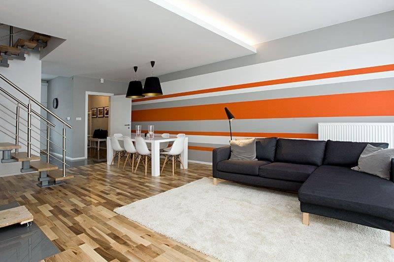 Wnętrze domu po remoncie: parkiet, biały dywan, szara sofa narożna, ściana w poziome pasy pomarańczowo-szare, biały stół z krzesłami, schody z metalową balustradą.