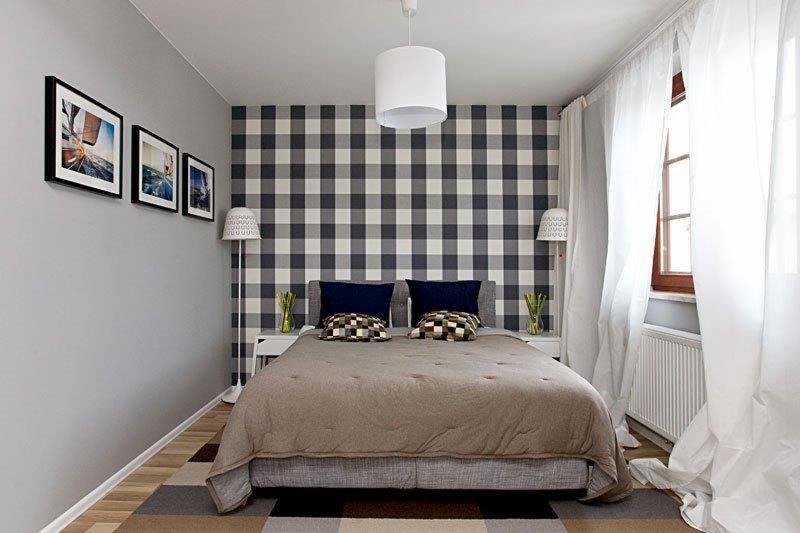 Sypialnia w domu w Wilanowie po remoncie: łóżko z szarą ramą i beżową narzutą, ściana w kratę, białe lampy podłogowe i wisząca, trzy obrazy na szarej ścianie, okno z białymi firankami.