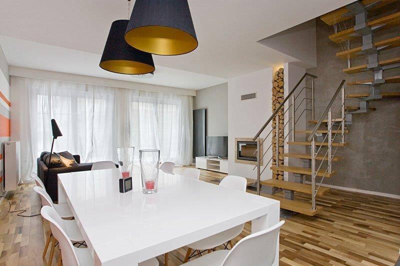 Nowoczesne wnętrze domu z białym stołem, krzesłami, drewnianymi schodami z metalową barierką, kominkiem i dekoracyjnym drewnem przy ścianie, oświetlone designerskimi lampami z czarnymi abażurami...