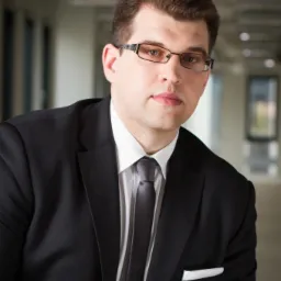 Portret mężczyzny w garniturze, okularach, z białą poszetką w kieszonce, na neutralnym tle biurowym.