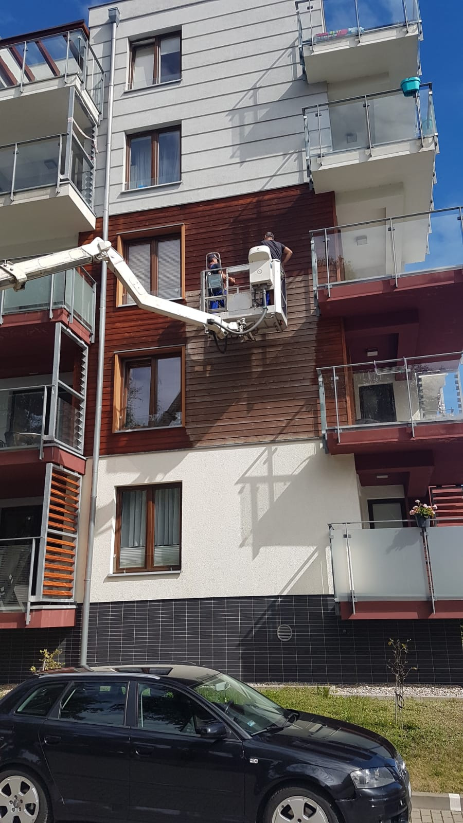 Dwóch pracowników na podnośniku koszowym czyści drewnianą elewację budynku wielorodzinnego, widoczny fragment czarnego samochodu na pierwszym planie.