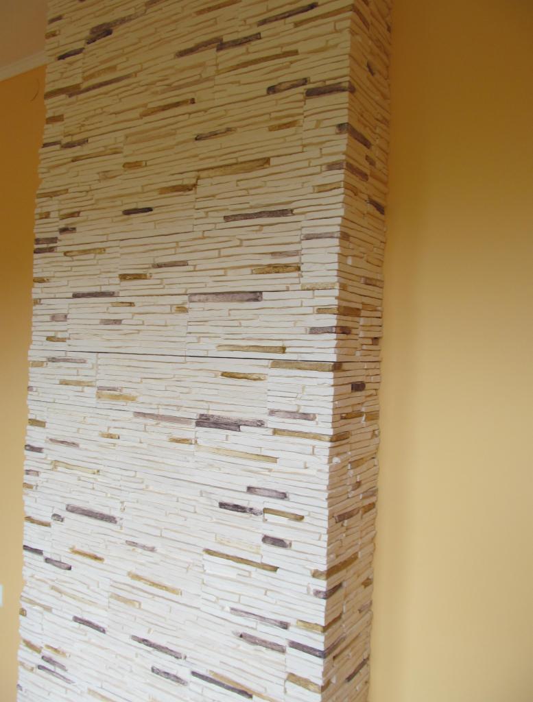 Dekoracyjny narożnik ściany wykończony okładziną z wąskich, poziomych płytek imitujących kamień w odcieniach beżu, brązu i szarości, na tle gładkiej, żółtej ściany.