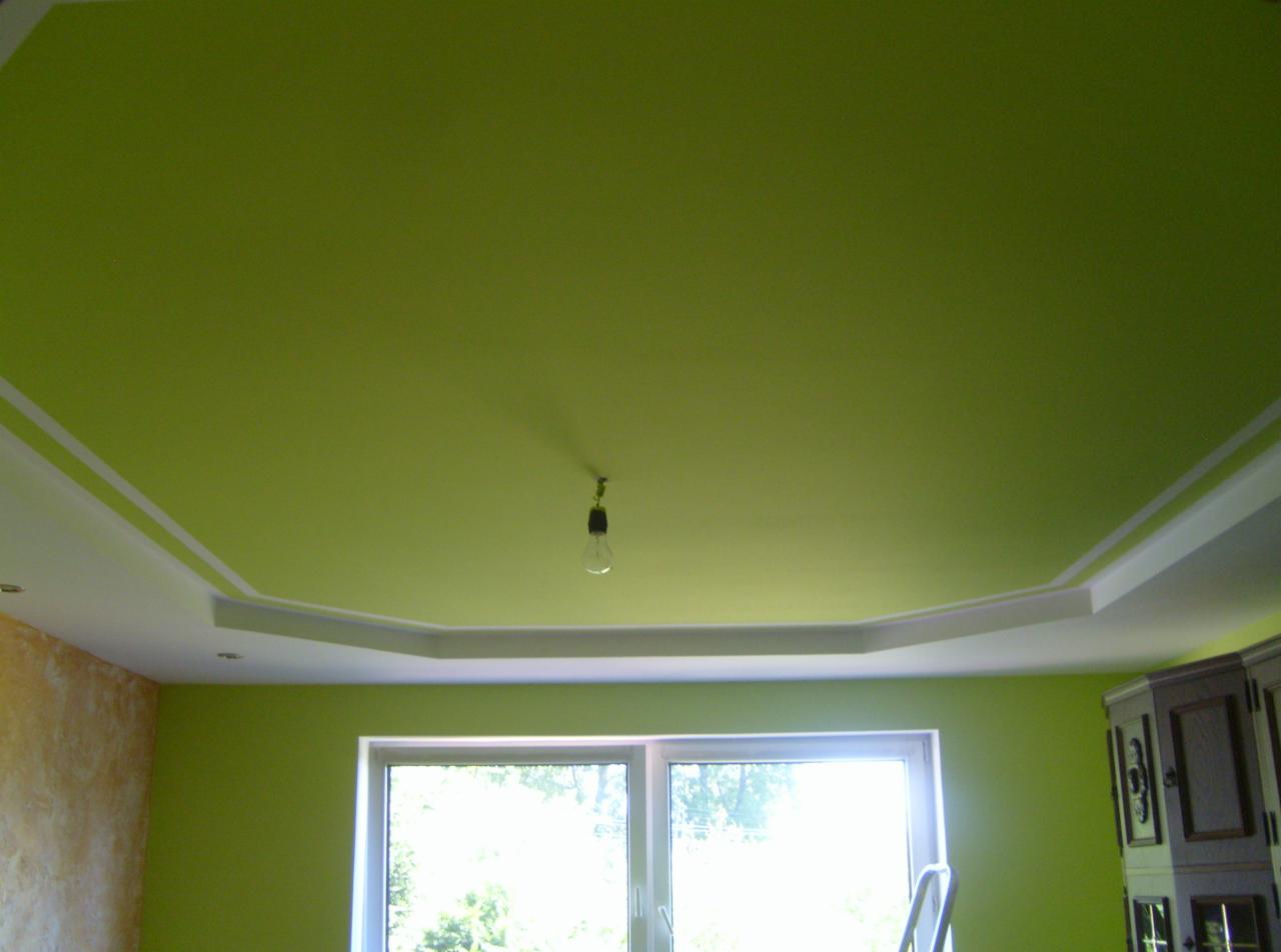 Pomieszczenie po malowaniu ścian i sufitu na kolor limonkowy, z białymi listwami dekoracyjnymi pod sufitem i wiszącą żarówką bez klosza, widok na okno i fragment ściany z fakturą.