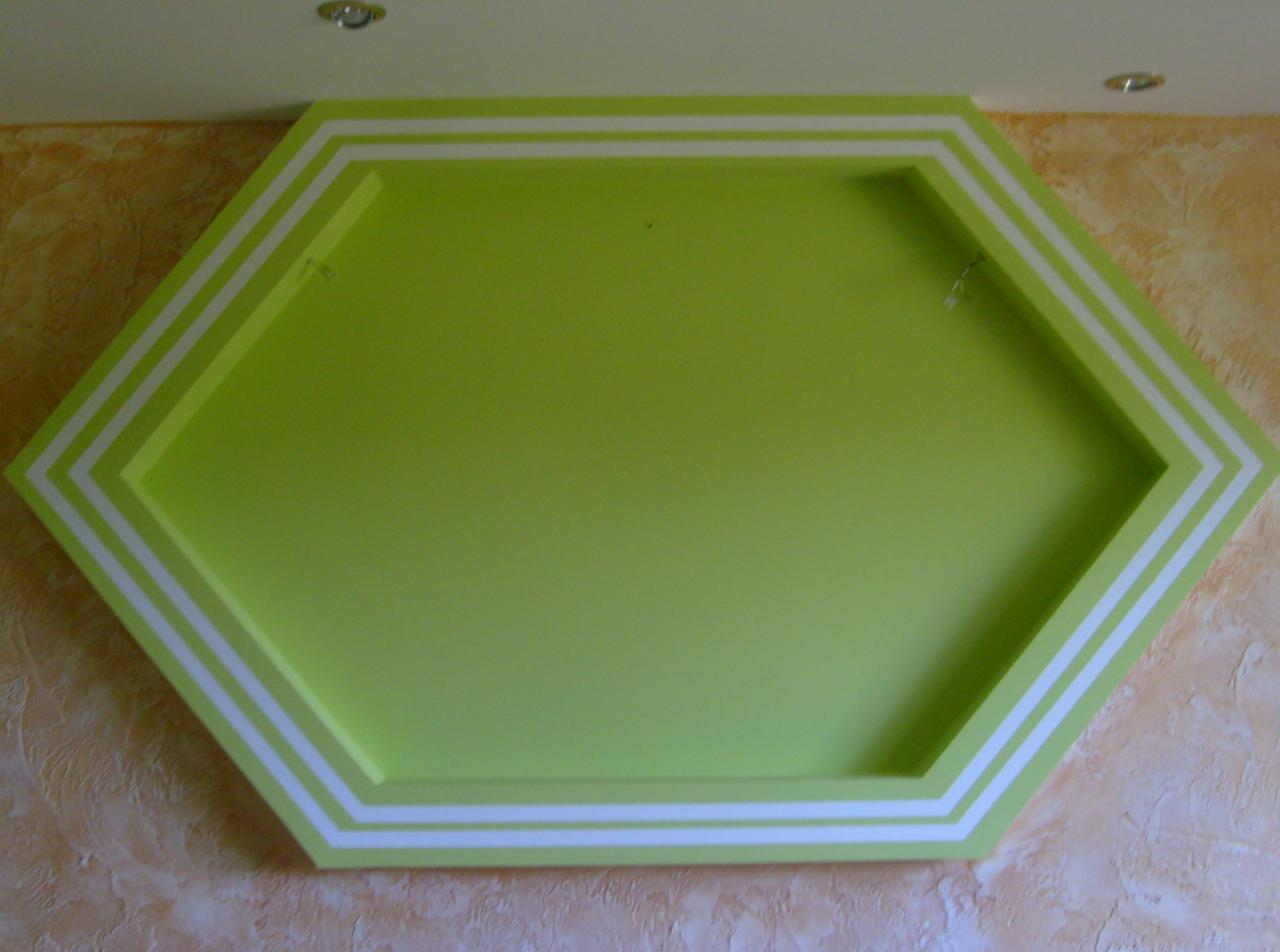 Sześciokątna, zielona dekoracja ścienna z białymi paskami na tle ściany o fakturze tynku.