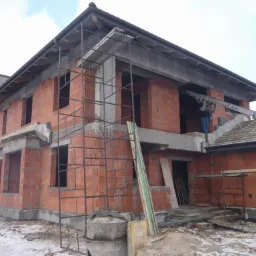 Dwukondygnacyjny dom murowany w stanie surowym otwartym, z widocznym rusztowaniem, elementami szalunków i częściowo ukończonym dachem, na placu budowy z resztkami śniegu.