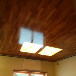 Drewniany sufit z parkietu w pomieszczeniu z oknem i grzejnikiem, widoczne odbicia światła od lamp.