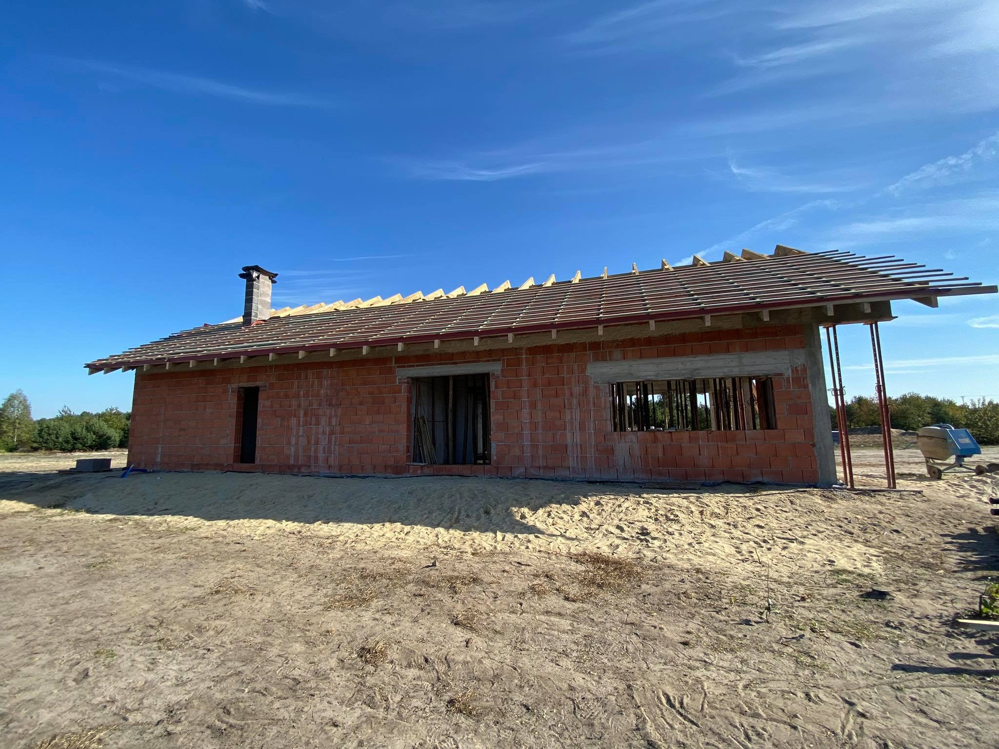 Budowa domu jednorodzinnego w stanie surowym otwartym: widoczna ceglana konstrukcja ścian, więźba dachowa z częściowo ułożoną dachówką ceramiczną oraz komin. Teren wokół budynku pokryty piaskiem.