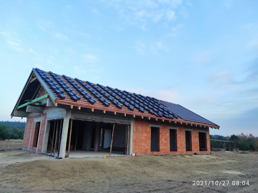 Budowa domu jednorodzinnego w trakcie, widoczna konstrukcja z cegły i częściowo ułożona dachówka ceramiczna w kolorze grafitowym na więźbie dachowej, tło stanowi las i błękitne niebo.