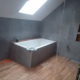 Wanna obudowana szarą płytką w łazience na poddaszu ze skośnym sufitem i oknem dachowym, pomalowane linie wyznaczają układ płytek na drewnopodobnej podłodze.