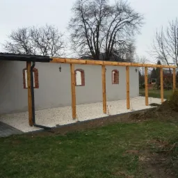 Zadaszenie tarasu z drewnianą konstrukcją, żwirowym podłożem i widocznymi elementami oświetlenia zewnętrznego przy budynku z oknami w kształcie łuków.