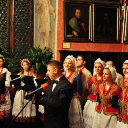 Występ chóru w strojach ludowych na tle zabytkowych obrazów i dekoracji ściennych. Dyrygent w smokingu z mikrofonem.