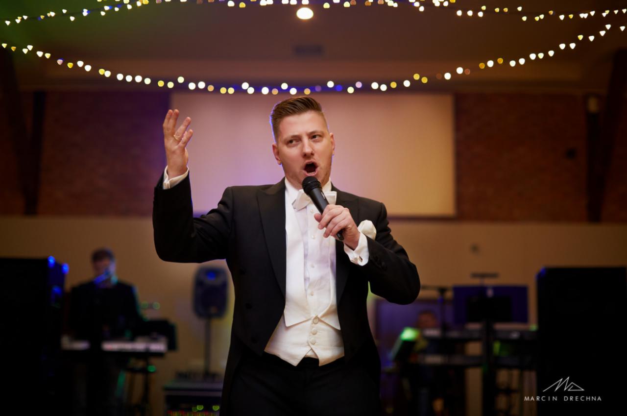 Mężczyzna w smokingu z mikrofonem w dłoni, gestykulujący podczas występu na tle dekoracji świetlnych i sprzętu muzycznego.