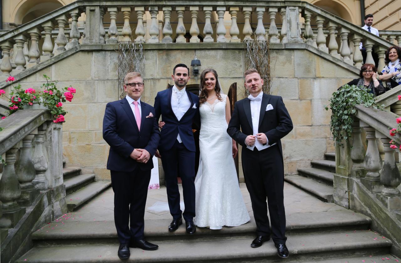 Para młoda z gośćmi pozuje na kamiennych schodach przed budynkiem z balustradami, różowe kwiaty w donicach i bluszcz zdobią schody.