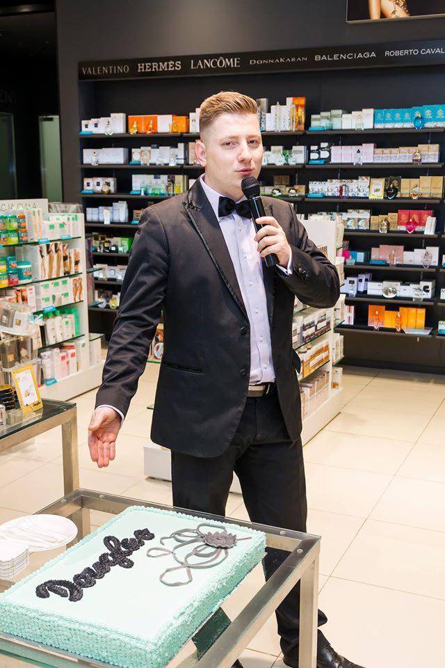 Mężczyzna w garniturze z mikrofonem prezentuje tort z napisem 'Douglas' na tle półek z kosmetykami.