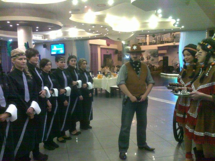 Obsługa eventu tematycznego: kelnerki w strojach stylizowanych na Indianki niosące napoje, kelnerzy w smokingach oraz mężczyzna przebrany za kowboja w kapeluszu i kamizelce na tle sali bankietowej.