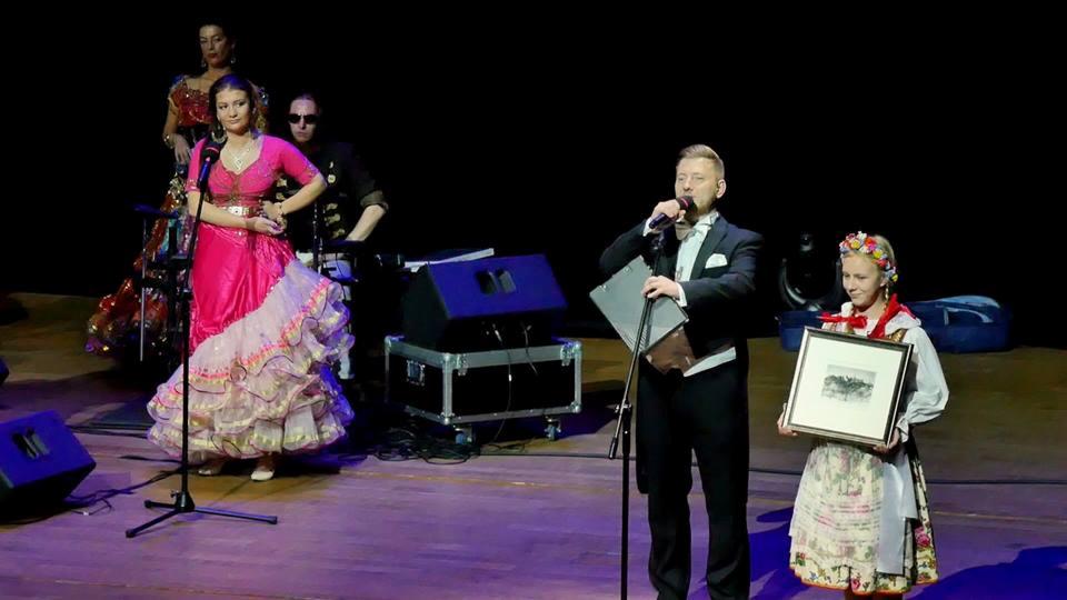 Scena z konferansjerem w smokingu, wręczającym nagrodę dziewczynie w stroju ludowym, obok tancerka w różowej sukni z falbanami i zespół muzyczny w tle.