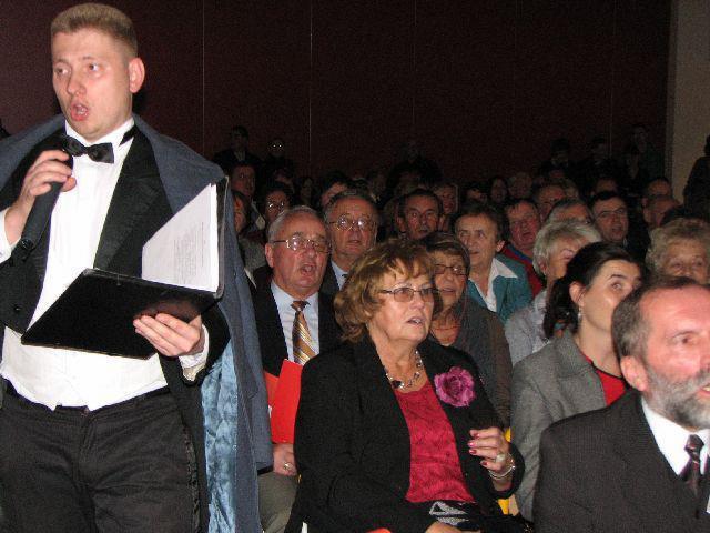 Mężczyzna w muszce i marynarce prowadzi śpiew chóru, trzymając czarny segregator z nutami przed zgromadzoną publicznością w ciemnym pomieszczeniu.