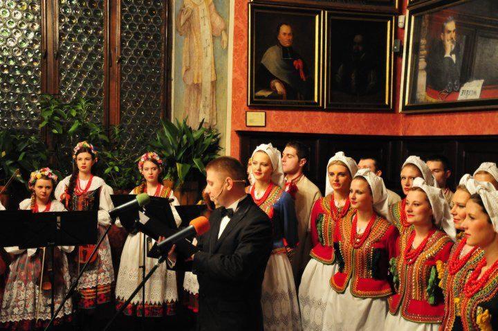 Występ chóru w strojach ludowych na tle zabytkowych obrazów i dekoracji ściennych. Dyrygent w smokingu z mikrofonem.