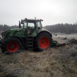 Zakład Usług Leśnych Mariusz Jakubaszek - Zielony traktor z czerwonymi kołami stoi na zroszonej łące na tle lasu w pochmurny dzień.