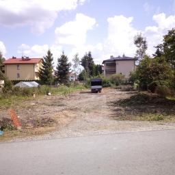 Zakład Usług Leśnych Mariusz Jakubaszek - Teren po wycince drzew z widocznymi pieńkami, gałęziami i małą przyczepką na tle zabudowy jednorodzinnej w słoneczny dzień.