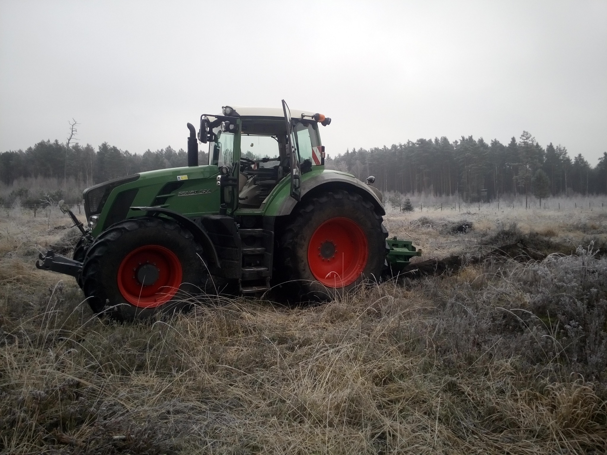 Zielony traktor z czerwonymi kołami stoi na zroszonej łące na tle lasu w pochmurny dzień.