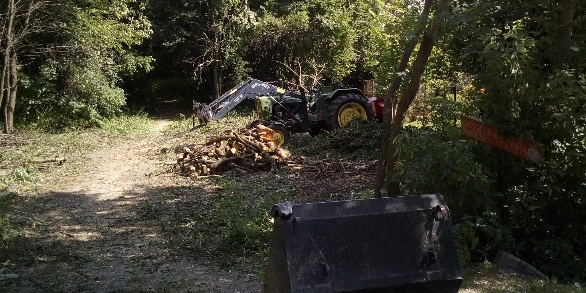 Zielony traktor z ładowarką WOL-MET zbiera pocięte gałęzie w lesie, w tle widoczny znak 'Sprzedam'.