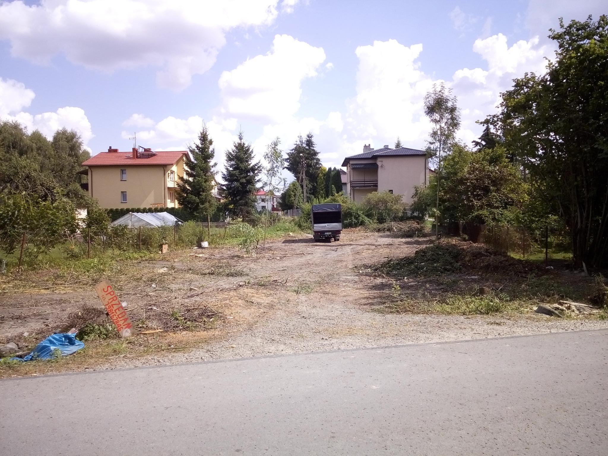 Teren po wycince drzew z widocznymi pieńkami, gałęziami i małą przyczepką na tle zabudowy jednorodzinnej w słoneczny dzień.