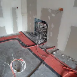 Instalacja ogrzewania podłogowego w surowym stanie, widoczne czerwone i czarne rury ułożone na podłodze, rozdzielacz w ścianie, ściany w trakcie szpachlowania.