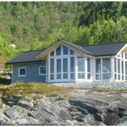 Tatran 3R Sp. z o.o. - Nowoczesny dom z dużymi oknami na skalistym podłożu, otoczony zielenią. Dach pokryty ciemną dachówką, elewacja w odcieniach szarości. Jasne, naturalne światło.