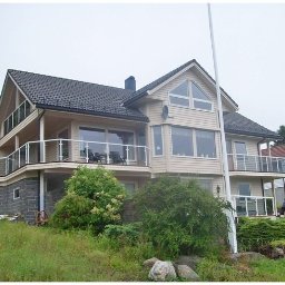 Tatran 3R Sp. z o.o. - Dwukondygnacyjny dom szkieletowy z balkonami i szklanymi balustradami, wykończony jasnym sidingiem, z ciemnym dachem i kamienną podmurówką, otoczony zielenią.