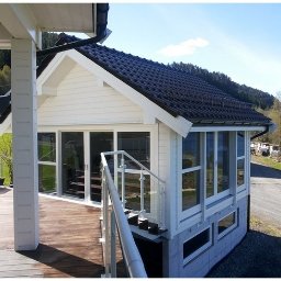 Tatran 3R Sp. z o.o. - Biały dom z ciemnym dachem, widok z boku z drewnianego tarasu. Widoczne okna, schody z metalową barierką i otoczenie z zielenią i drogą.