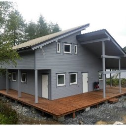 Tatran 3R Sp. z o.o. - Nowoczesny dom szkieletowy z szarą elewacją i drewnianym tarasem, otoczony zielenią i jeziorem w tle. Charakterystyczny asymetryczny dach i minimalistyczne okna.