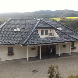 Tatran 3R Sp. z o.o. - Biały dom z ciemną dachówką na tle zielonych wzgórz. Widoczne okna dachowe i dekoracje kwiatowe. Na podjeździe stoi mężczyzna.