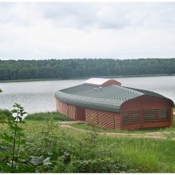 Tatran 3R Sp. z o.o. - Nietypowy, drewniany budynek z zaokrąglonym dachem, położony nad jeziorem, otoczony zielenią. Widoczne elementy konstrukcyjne i naturalne otoczenie.