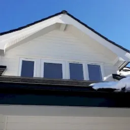 Fragment białego domu z czarnym dachem, widoczne trzy okna na poddaszu i resztki śniegu na dachu, błękitne niebo w tle, logo firmy w prawym dolnym rogu.