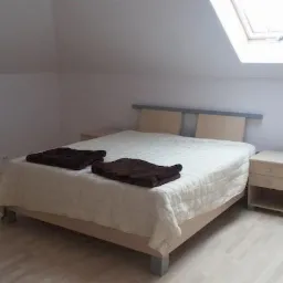 Jasna sypialnia na poddaszu z oknem dachowym, łóżkiem z beżową ramą i dwiema brązowymi narzutami, drewniana podłoga, minimalistyczny wystrój.