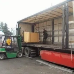 Wózek widłowy podnosi drewniane elementy na platformę ciężarówki; w tle widoczny pracownik zabezpieczający ładunek.