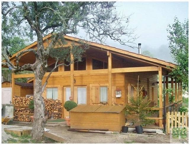 Drewniany dom z bali z widocznym stosem drewna obok, otoczony zielenią i drzewami, w tle mglista sceneria. Widoczny fragment werandy i mały ogródek.
