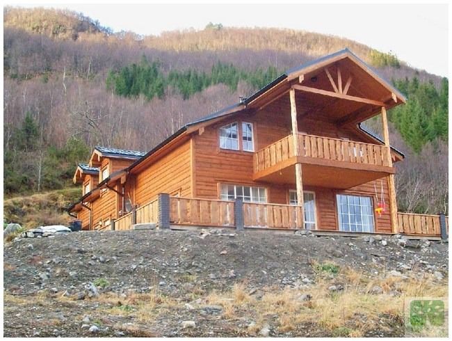 Drewniany dom z balkonem na tle zalesionego wzgórza. Widoczne detale konstrukcyjne i naturalna kolorystyka drewna. Ujęcie z dołu podkreśla bryłę budynku.