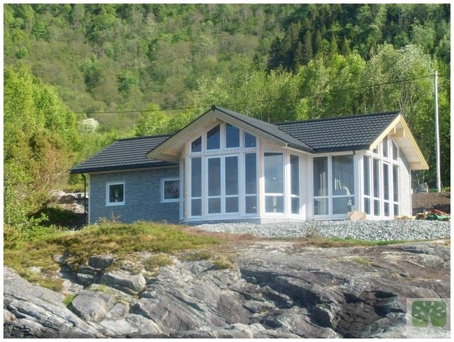 Nowoczesny dom z dużymi oknami na skalistym podłożu, otoczony zielenią. Dach pokryty ciemną dachówką, elewacja w odcieniach szarości. Jasne, naturalne światło.