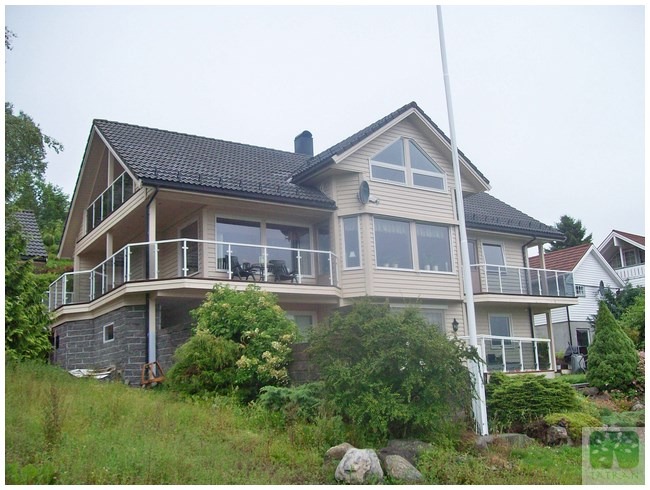 Dwukondygnacyjny dom szkieletowy z balkonami i szklanymi balustradami, wykończony jasnym sidingiem, z ciemnym dachem i kamienną podmurówką, otoczony zielenią.