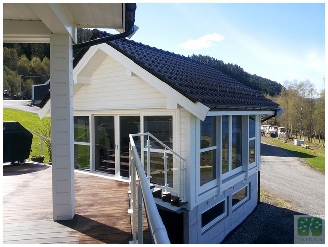 Biały dom z ciemnym dachem, widok z boku z drewnianego tarasu. Widoczne okna, schody z metalową barierką i otoczenie z zielenią i drogą.