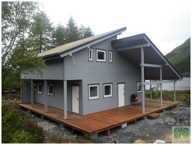 Nowoczesny dom szkieletowy z szarą elewacją i drewnianym tarasem, otoczony zielenią i jeziorem w tle. Charakterystyczny asymetryczny dach i minimalistyczne okna.