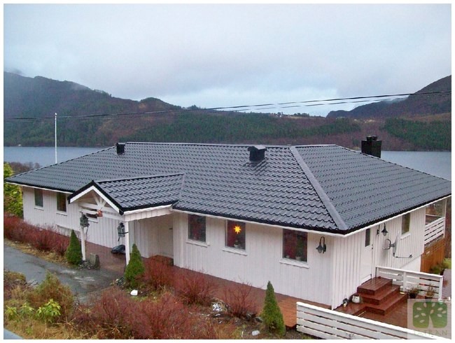 Biały dom z ciemnoszarym dachem na tle jeziora i zalesionych wzgórz. Widok z góry ukazuje dach z blachy trapezowej i fragment otoczenia.