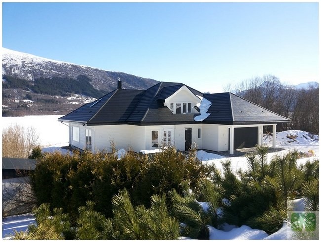 Biały dom z garażem i ciemnym dachem na tle gór pokrytych śniegiem. Część dachu pokryta śniegiem. W tle błękitne niebo i zimowy krajobraz.