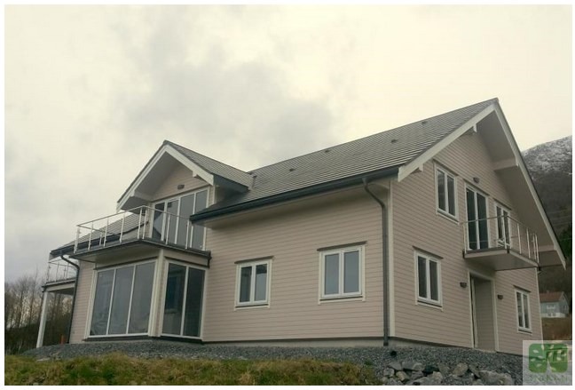 Jasny, dwukondygnacyjny dom z balkonami i dużymi oknami, wykończony drewnianą elewacją. Widok z dołu na tle pochmurnego nieba i wzgórz.